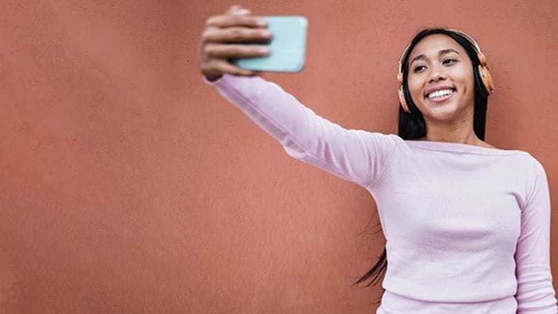 A young Black woman wearing headphones is smiling and taking a selfie with her smartphone against a terra cotta colored wall.