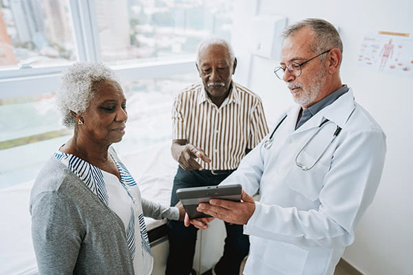 An older adult couple is looking at a digital tablet with their health care professional in a medical office.